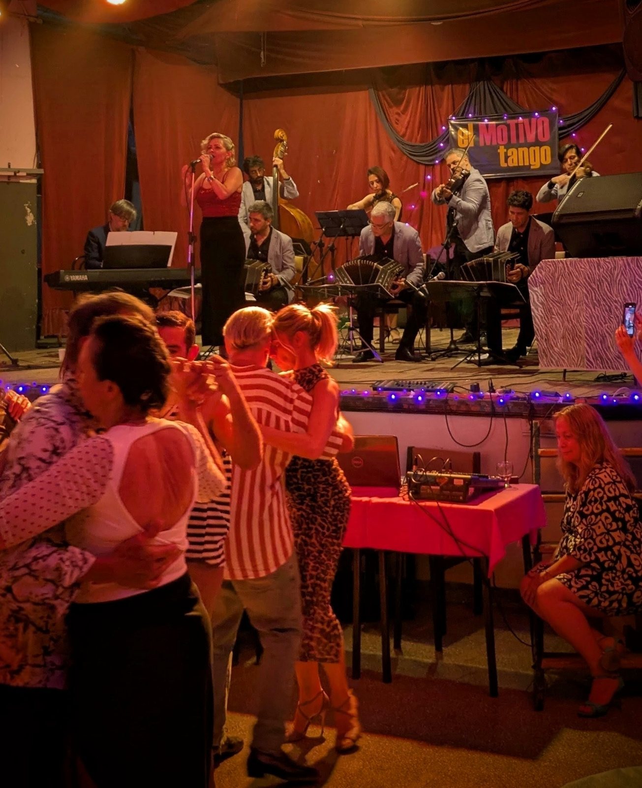 A milonga on an orchestra night, seen from a table: couples on the floor, the orchestra on the stage behind